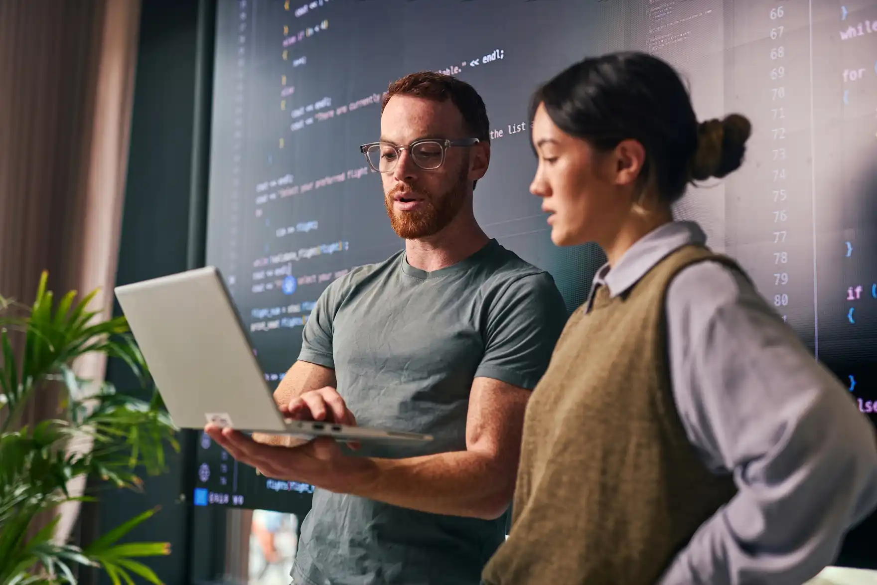 Two developers reviewing work on a laptop in front of a wall-sized display of code